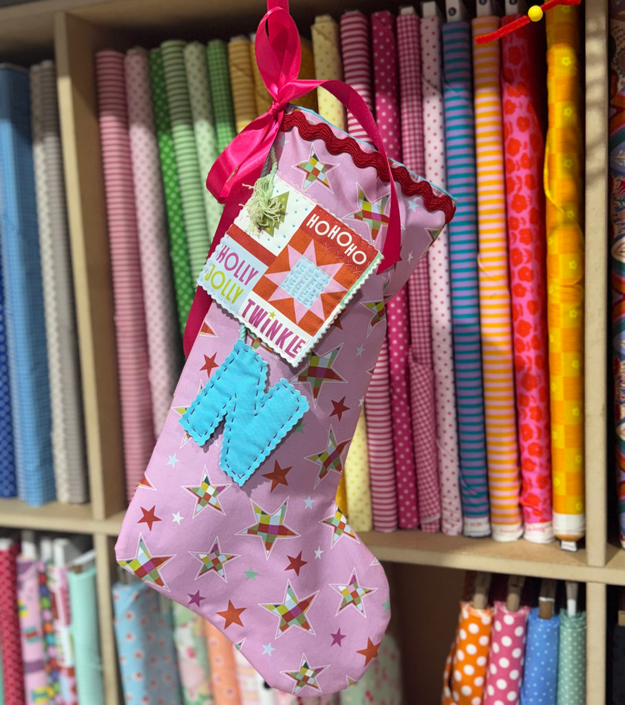 Pink stocking with colorful patches hanging in front of a shelf of fabric rolls.